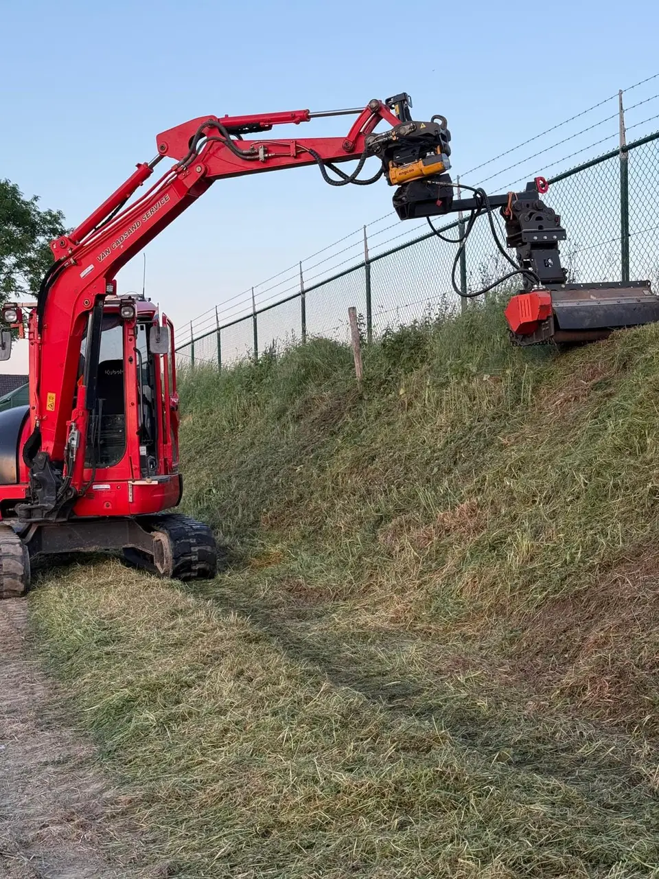 Kraan met klepelmaaier aan het werk op een steile berm langs een hek
